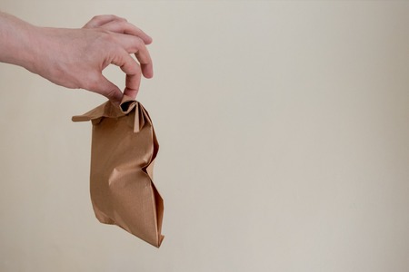 A man holds a brown paper bag with two fingers in his hand. Delivery or recycling concept. Copy space. Beige background.の写真素材