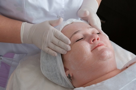 Therapeutic and cosmetological facial massage of a fat woman in the health and beauty salon.The hands of a professional beautician in white medical gloves smooth the skin face of a smiling girl.の写真素材
