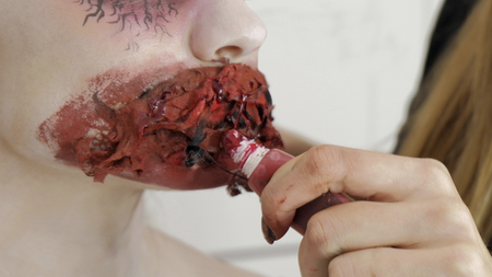 Preparing for Halloween or for a photo shoot. Makeup artist makes a bloody mouth make-up with red lip gloss close-up.の写真素材