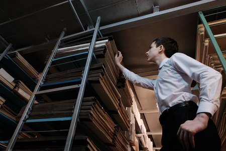 A teenager in a white shirt pulls out a book from the rack in a book depository or archive room. Schoolboy studying historical documents. The concept of a historical library, back to school or popularization of reading.の写真素材