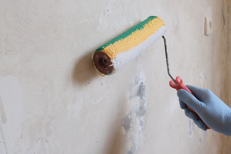 A male blue-gloved hand primed a concrete wall with a roller. The concept of home repair, finishing works, preparing walls for plastering and painting. Copy space.の写真素材