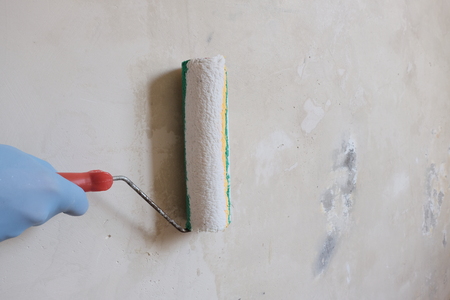 A hand in a blue glove holds a roller for finishing work. The process of priming the concrete wall. The concept of repairing the premises, construction work, preparing the room for painting.Copy space.の写真素材