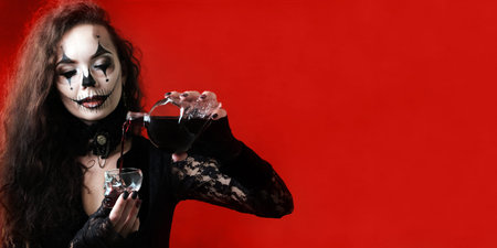 A Halloween-style brunette wearing black clothes pours a red liquid that looks like blood into a...