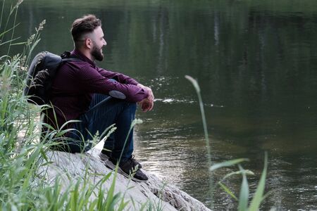 Joyful stylish guy with a beard. Traveler with a backpack in jeans and shirt is sitting on a stone by the river and looking ahead. The concept of human unity with nature, travel and lifestyle.の写真素材