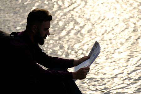 Silhouette traveler with a map in his hands. A guy with a beard in a shirt is sitting at sunset by a river, lake or sea. The concept of human unity with nature, travel and lifestyle. Copy space.の写真素材