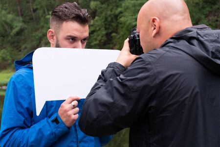 Photosession in nature. Bald photographer works with a model guy. A young man hides his face behind a white sign.の写真素材