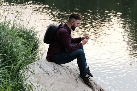 Traveler with a backpack and a smartphone. A bearded guy in jeans and a T-shirt with a phone in his hands sits on a stone by the river. Concept of travel, tourism and lifestyle.の写真素材
