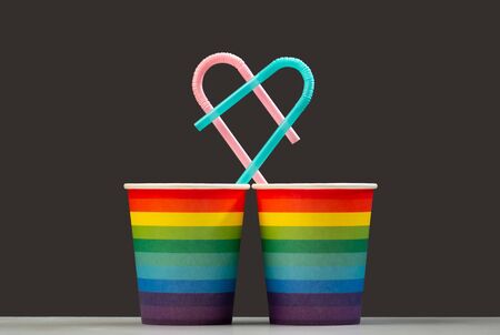 Together forever. Two LGBT color paper cups with pink and blue heart shaped cocktail tubes. The concept of unification and love of people of homosexuality or a symbol of the holiday and anniversary of living together. Gray and rainbow background. Isolateの写真素材