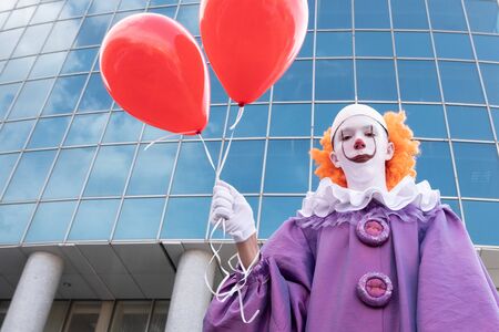 It is against the backdrop of a tall glass building. Scary clown with red balls in the city. Costume for carnival or for halloween.の写真素材