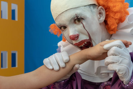 A teenage boy in the image of a clown with sharp teeth pretends to bite someone's hand. Street...