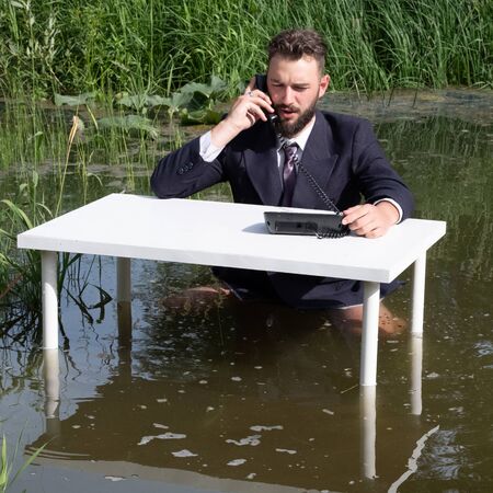 Businessman without pants in a swamp. A man with a beard in a suit as a clerk or office manager sits in a quagmire at a white table and talks on the phone. Concept of crisis or routine tech support.の写真素材