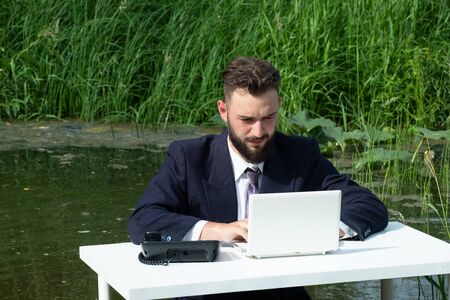 Office in the swamp. A young businessman with a beard in a suit as a manager or clerk sits in a quagmire at a white table and works on a laptop. Concept of freelance, crisis or routine.の写真素材