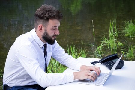Young brutal guy with a beard in the clothes of an office worker or manager sits at a table and works on a laptop against the background of the river. Concept of businessman routine or freelancer job. Dream of vacation.の写真素材