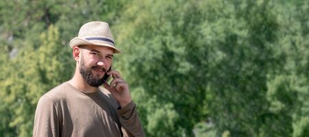 A bearded guy in a hat speaks on the phone and looks sadly into the frame. The lack of telephone communications in nature. The concept of tourism, leisure and travel, a technical problem. Copy space. Template for a banner.の写真素材