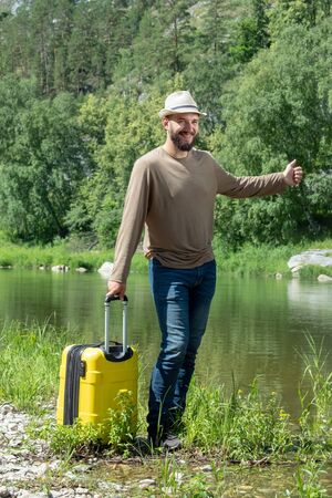 Bearded guy in a hat smiles. A traveler with a yellow suitcase on the shore of a mountain river is waiting for a taxi. A curious case. Tourism concept, thumb up, leisure and travel, transport problem.の写真素材