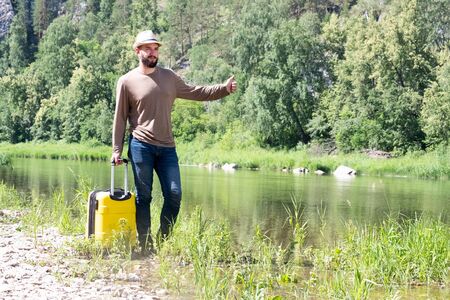 The traveler in the hat raised his hand and thumb up as if he were catching a taxi. Sad hipster young guy hitchhiking along a mountain river with a yellow suitcase. Copy space.の写真素材