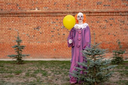 A guy in a clown or mime costume with a yellow ball near a small green Christmas tree against a brick wall for All Saints Day holiday. Street cosplay for halloween, carnival or party.の写真素材