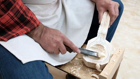 An elderly man cuts a spoon out of a wooden block with a sharp chisel. Men's hands with a tool closeup. Traditional handicraft woodworking or hobby.の写真素材
