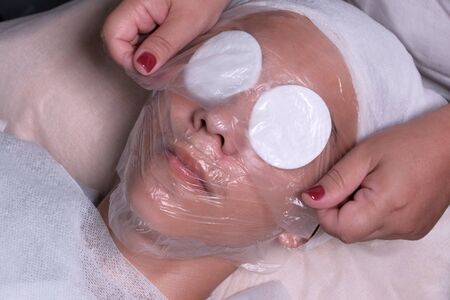 Teenage cosmetology procedure. Hands of a beautician cover the face of a boy of 15 years with a transparent film. The guy lies with cotton pads in front of his eyes. Health and skin care in a cosmetology center or clinic.の写真素材