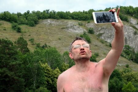 An unshaven middle-aged man takes off taking a selfie against the backdrop of the mountains. Bespectacled photographer without clothes on nature holds the phone in his left hand. Funny face while relaxing, vacation and traveler hobby.の写真素材