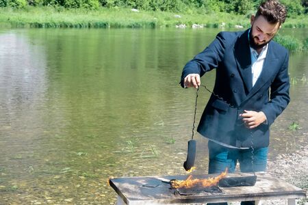 Businessman burned office equipment outdoors during the vacation. The concept of hateful work. Destruction of digital data or non-compliance with safety precautions. A laptop and a phone burned out on a table near the river. The male hand of the manager holds the wire the telephone receiver on fire.の写真素材