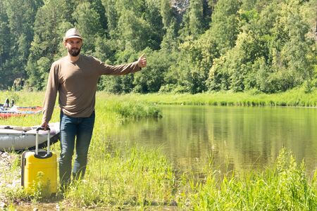 Hipster young man hitchhiking along a mountain river with a yellow suitcase. Travel and vacation. Summer or tropics. Copy space.の写真素材
