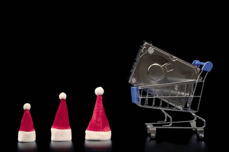 A laptop hard drive in a blue shopping basket and three red Santa Claus caps in line. Christmas sale and family discount concept, black friday, cyber monday. Template for online store advertising. Copy space. Black background.の写真素材