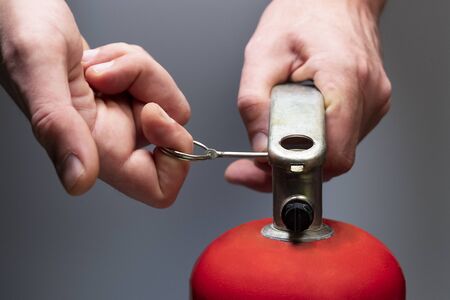 Actions in case of fire. Pull the safety pin out of the fire extinguisher by the ring. Mens hands close-up. Safety measures in the office and in the workplace. Emergency Prevention. Grey background. Shallow depth of field. Selective focus.の写真素材