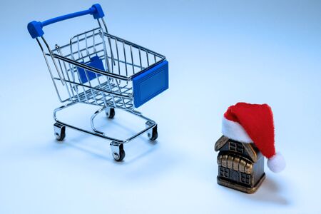 Souvenir house with santa claus hat and stroller for shopping. The concept of housewarming, New Years mortgage, Christmas work of a realtor. Discounts, promotions and sale of real estate. Blue tinted background.の写真素材