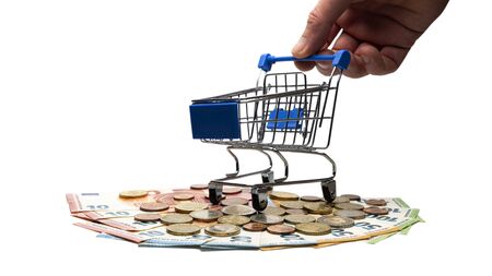 The male hand holds a shopping trolley, which stands on banknotes and euro coins. Close-up. Isolated. The concept of the consumer basket in European countries, crisis or wealth, sale or accumulation.の写真素材