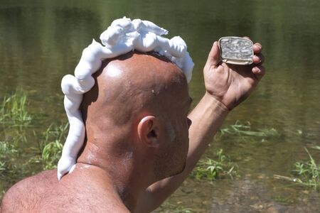 A bald guy with a shaving foam mohawk looks in the mirror. Ecotourism or traveler lifestyle. A man shaves in nature near a river with green grass.の写真素材