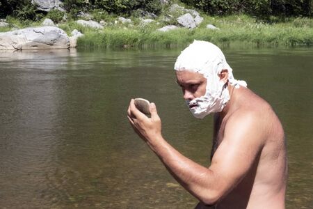 The head and face of a man are covered with soap foam. A guy with a wild look looks at himself in the mirror. A tourist shaving in the mountains or a traveler lifestyle near the river. The concept of hygiene in nature.の写真素材