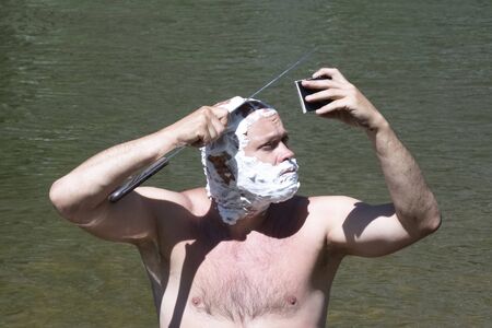 A brutal guy looks at himself in the mirror and shaves his bald head with a sharp machete knife on a background of water. The head and face of a man are covered with soap foam. Traveler lifestyle. The concept of hygiene in nature.の写真素材