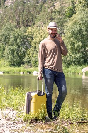 A guy in a hat and jeans is talking on the phone outdoors. A concept of leisure and travel. Hipster young man hitchhiking along a mountain river with a yellow suitcase.の写真素材