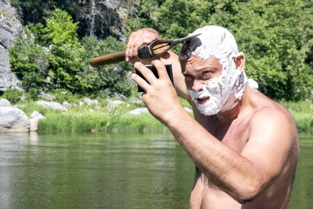 A brutal Caucasian guy shaves his bald head with a sharp ax at himself in the mirror. The head and face of a man are covered with soap foam. Traveler lifestyle in the mountains. The concept of hygiene in nature.の写真素材