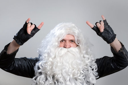 A tough guy in a wig and a beard like Santa Claus makes a heavy metal gesture. Man in a black biker jacket and gloves as rock fan or heavy metal musician looks at the camera.の写真素材