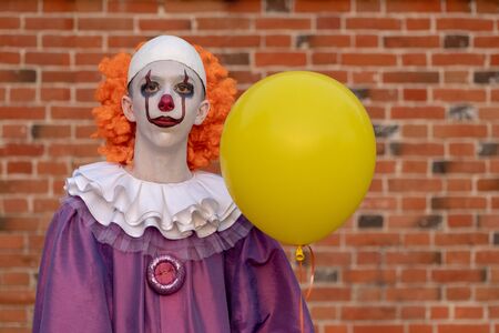 A guy in a clown costume with a yellow balloon against a brick wall. Costume for carnival, all saints day or halloween.の写真素材