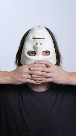 A long-haired man holds a white mask upside down near his face. Vertical frame. The concept of hiding a person, peeping, mental problems, a split personality, psychedelia or voodoo ritual.の写真素材