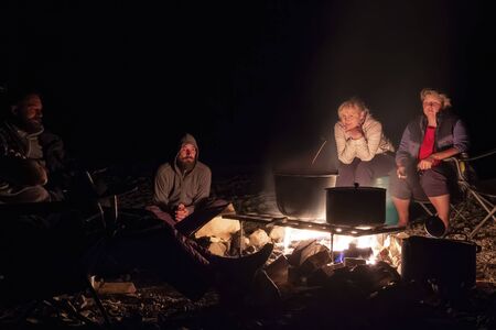 Group of backpackers relaxing near campfire after a hard day. Two women and two men look at the fire on which food is cooked, tourist background. Dark night. Copy space.の写真素材