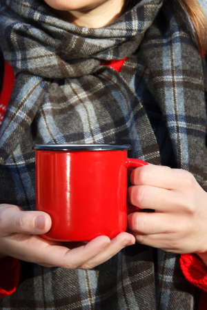 girl in a cashmere scarf in a cage holds a red mug with tea in her palmsの写真素材