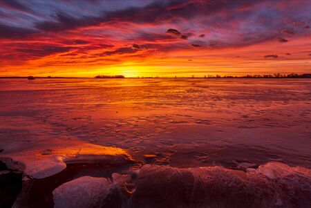 The sun lights up the clouds at sunrise or sunset above the frozen winter lake with rocks in the foregroundの写真素材