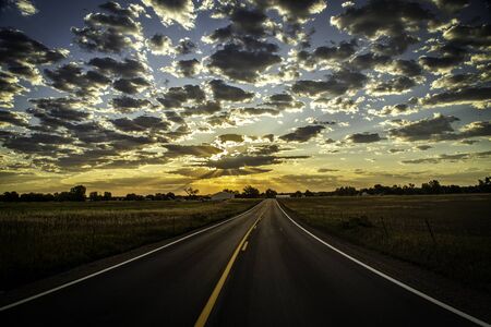 A country Paved Road leading into the Horizon as sun rays beam out of the clouds at sunrise or sunsetの写真素材