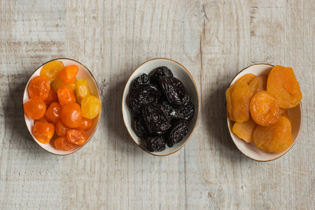 Three plates with dried fruits on wooden background: dried mandarins, prunes and dried apricots.