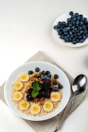 Oatmeal porridge with bananas, walnuts, blackberries, blueberries, honey and mint in a white bowl on a white background. Healthy breakfast and homemade diet foodの写真素材