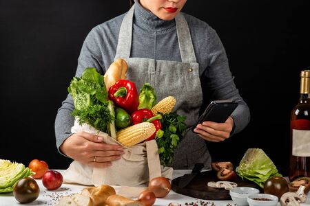 Order food online. Woman in the kitchen holds an eco fabric bag full of fresh organic vegetables and a smartphone in hand. Purchases concept. Cooking healthy foodの写真素材