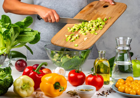 Woman adds chopped avocado to salad in a glass bowl, cooking in the kitchen. Healthy diet concept
