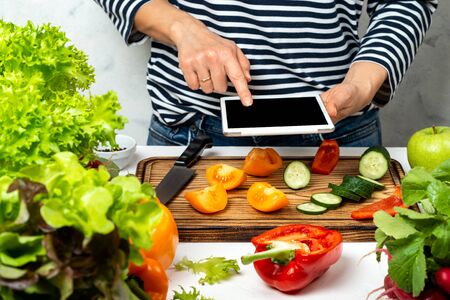 Woman cooking and holding tablet computer with blank screen in hand. Cutting vegetable ingredients and following recipe from the internet.の写真素材