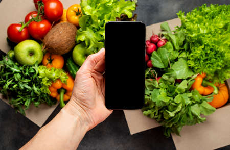 Female hand with smartphone and full boxes of different fresh organic vegetables. Delivery or Order food online concept.の写真素材