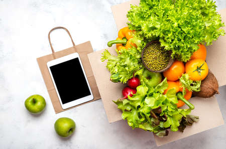 Box full of fresh organic products, tablet computer with blank screen and paper shopping bag. Delivery order food concept. Top view.の写真素材