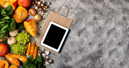 Online grocery shop concept. Tablet computer and shopping paper bag with pieces of pumpkin Different vegetables and greens. Banner Top view.の写真素材